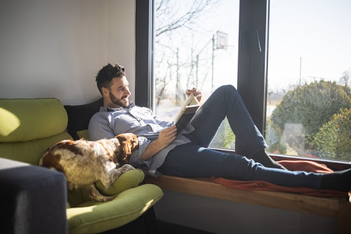 Young bearded man lying on wooden bench next to window and reading book while his dog lying next to him and napping