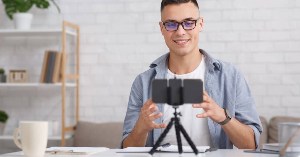 a man sits at a desk smiling into a smartphone on a tripod