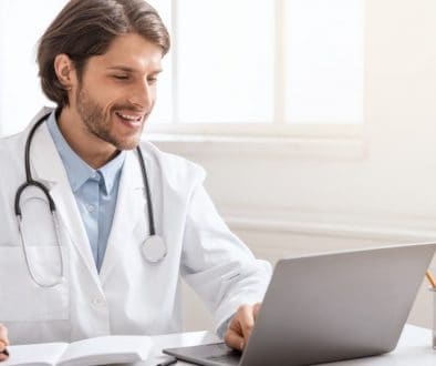 young male doctor in a lab coat with stethoscope over his shoulders smiles into laptop while writing notes
